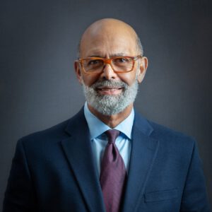 Professional headshot of Dr. Lomax in business suit with orange-framed glasses